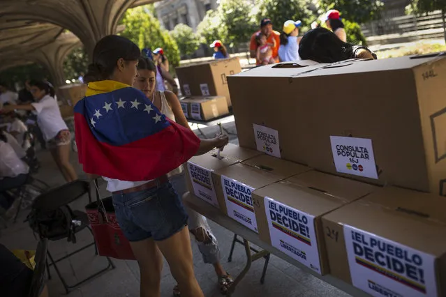 A woman, with a Venezuela flag over her shoulders, casts her ballot during a symbolic referendum in Madrid, Sunday, July 16, 2017. Venezuela's opposition called for a massive turnout Sunday in a symbolic rejection of President Nicolas Maduro's plan to rewrite the constitution, a proposal that's escalating tensions in a nation stricken by widespread shortages and more than 100 days of anti-government protests. White sheets on the boxes read in Spanish: “Popular referendum” and “People decide!”. (Photo by Francisco Seco/AP Photo)