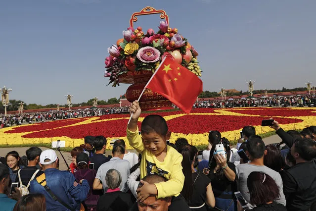 A Chinese boy waves a national flag on his father's shoulder as visitors gather near a giant basket decorated with replicas of flowers and fruits on display at Tiananmen Square on China's National Day in Beijing, Sunday, October 1, 2017. Hundreds of thousands foreign and domestic tourists flock to the square to celebrate the 68th National Day and the Mid-Autumn Festival over the week-long holidays. (Photo by Andy Wong/AP Photo)