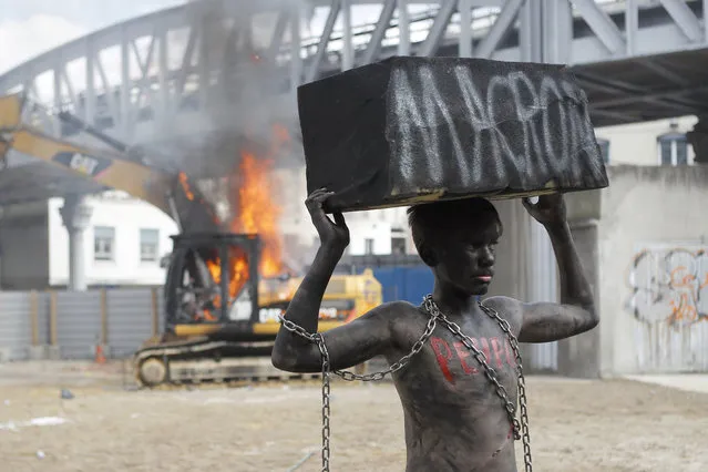 A chained activist, with his body painted in black, lifts a fake stone bearing the name of French President Emmanuel Macron and the word “people” on his torso as he poses for photographers while a digger set ablaze by demonstrators is seen on background during the traditional May Day rally in the center of Paris, France, Tuesday, May 1, 2018. Each year, people around the world take to the streets to mark International Workers' Day, or May Day. (Photo by Francois Mori/AP Photo)