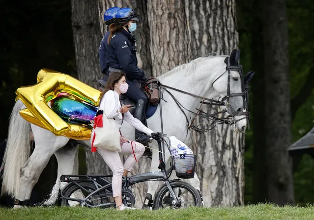 Police patrol as a woman rides her bike carrying balloons at the Bois de la Cambre park, during a party called “La Boum 2”, in Brussels, Saturday, May 1, 2021. Police put on extra patrols Saturday to monitor the gathering which is being held in defiance of Belgium's current COVID-19 regulations. (Photo by Olivier Matthys/AP Photo)