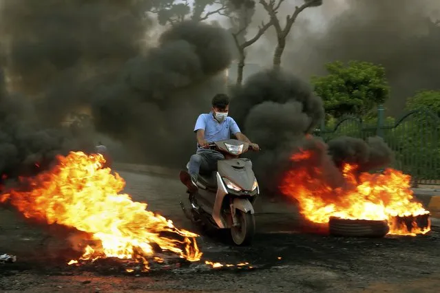 A man rides his scooter through the burning, that were set on fire by protesters to block a road, in Beirut, Lebanon, Thursday, June 24, 2021. Dozens of angry protesters, angered by deteriorating living conditions and government inaction, partially blocked Beirut's main highway to the capital's only airport, turning trash bin over and setting tires on fire. (Photo by Bilal Hussein/AP Photo)