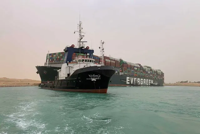 In this photo released by the Suez Canal Authority, a boat navigates in front of a massive cargo ship, named the Ever Green, rear, sits grounded Wednesday, March 24, 2021, after it turned sideways in Egypt’s Suez Canal, blocking traffic in a crucial East-West waterway for global shipping. An Egyptian official warned Wednesday it could take at least two days to clear the ship. (Photo by Suez Canal Authority via AP Photo)
