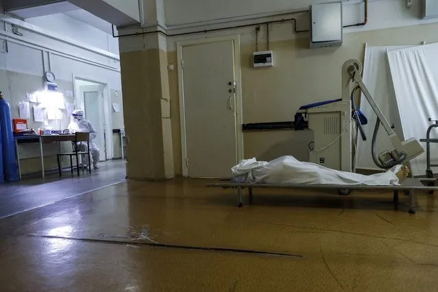 A medic wearing a special suit to protect against coronavirus fills documents as a body of a COVID-19 victim lies on a stretcher at an ICU of a hospital in Volgograd, Russia, Sunday, November 21, 2021. The latest surge in deaths comes amid low vaccination rates and lax public attitudes in Russia toward taking precautions. About 40% of Russia's nearly 146 million people have been fully vaccinated, even though the country approved a domestically developed COVID-19 vaccine – Sputnik V – months before most of the world. (Photo by Alexandr Kulikov/AP Photo)