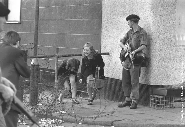 1969:  Two young women climbing under a barricade next to an armed soldier on the Falls Road, Belfast