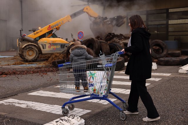 A woman, with her child, pushes a shopping trolley past a supermarket blocked by French farmers who dump tires and slurry as part of their protest over price pressures, taxes and green regulation, grievances shared by farmers across Europe, in Bon-Encontre near Agen, south of France, on January 25, 2024. (Photo by Nacho Doce/Reuters)