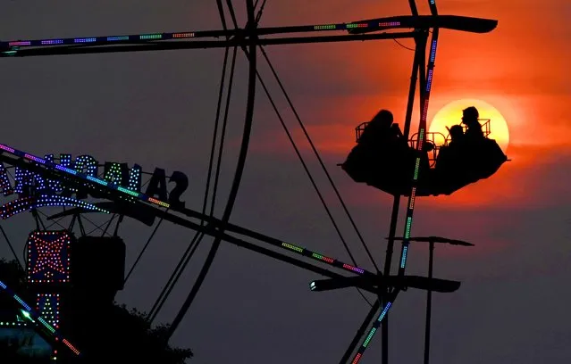 Ferris wheel riders are silhouetted against the rich setting sun in Gaithersburg, Maryland on May 22, 2023. Wildfires in Canada have caused more colorful sunsets recently due to smoke in the atmosphere. The more dramatic than usual sunsets will continue as the fires are still burning. The Jolly Shows carnival will remain set up in the parking lot of the shuttered Lake Forest Mall through May 28th. (Photo by Michael S. Williamson/The Washington Post)