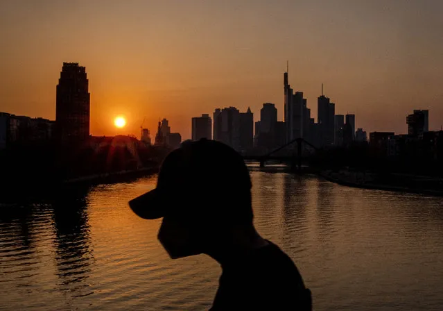 A man wears a face mask as he walks on a bridge over the river Main as the sun sets in Frankfurt, Germany, Thursday, March 24, 2022. (Photo by Michael Probst/AP Photo)