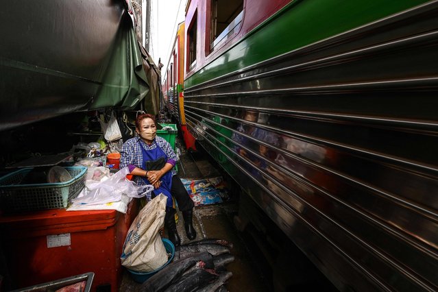 This photograph taken on June 4, 2022 shows fish vendor Somporn Thathom sitting at her stall as a passenger train passes through the Mae Klong railway market in Samut Songkhram province, around 80 kms (50 miles) southwest of Bangkok. Six times a day at the market, local customers and foreign tourists scramble into nooks and crannies while vendors calmly move their woven baskets of goods away from the tracks and close their umbrellas. (Photo by Manan Vatsyayana/AFP Photo)