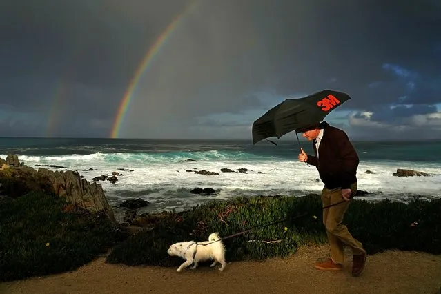 A man huddles under an umbrella as he walks a dog along the shoreline at Pacific Grove, Calif., Friday, February 2, 2024, with a rainbow in the background. (Photo by Rich Pedroncelli/AP Photo)