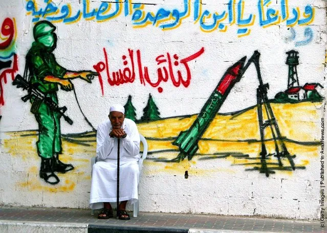 An elderly Palestinian man sits in front of a wall with a mural showing a masked militant from Ezz El-Deen Al-Qassam Martyrs' Brigade, the military wing of the Islamic resistance movement Hamas, as he fires a homemade Qassam rocket towards Israeli settlements, in Jabaliya refugee camp northern Gaza Strip August 28, 2003