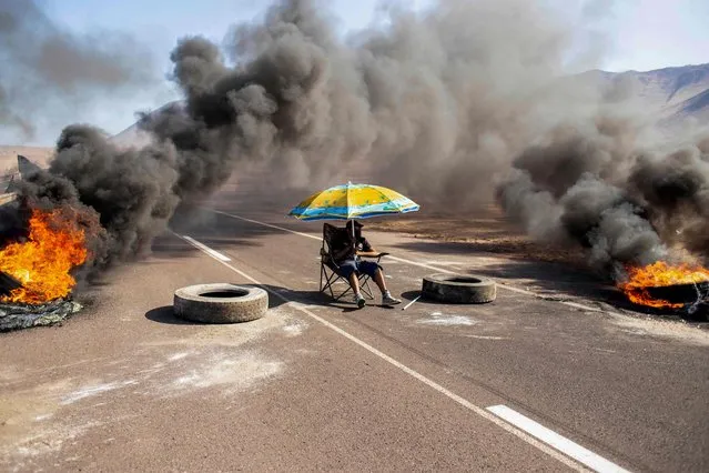 A demonstrator sits under an umbrella while blocking an access route to Iquique, Chile, during a regional strike called by different organizations against illegal immigration, on January 31, 2022. The city of Iquique, in the north of Chile and near the border with Bolivia, experienced a second day of agitation Monday with blockades at its entrance roads and the closure of its airport, in protest against the increase in crime that some associate with the illegal immigration. (Photo by Diego Reyes/AFP Photo)