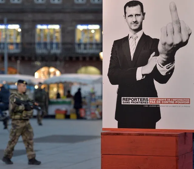 A French soldier walks past a poster by non-profit organisation Reporters Without Borders (Reporters sans frontières) of Syrian President Bashar al-Assad showing his middle finger, as thousands of people gather at the central square in Strasbourg, eastern France, on November 18, 2015, to pay tribute to the victims of the attacks of November 13. Gunmen and suicide bombers went on a killing spree in Paris on November 13, attacking a concert hall, bars, restaurants and the Stade de France. Islamic State jihadists operating out of Iraq and Syria released a statement claiming responsibility for the coordinated attacks that killed 129 people and left 352 others injured. (Photo by Patrick Hertzog/AFP Photo)