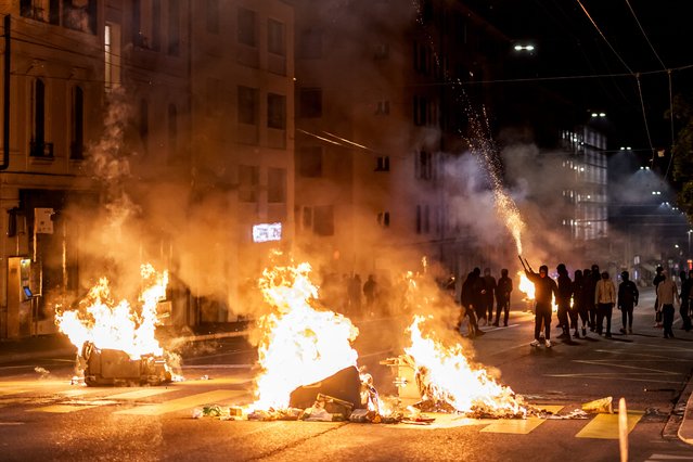 Young people wearing face coverings launch fireworks from behind fires in a street in Lausanne, on August 25, 2025, during a second night of rioting following the death of a teenager who tried to escape local police while driving a stolen scooter. (Photo by Fabrice Coffrini/AFP Photo)