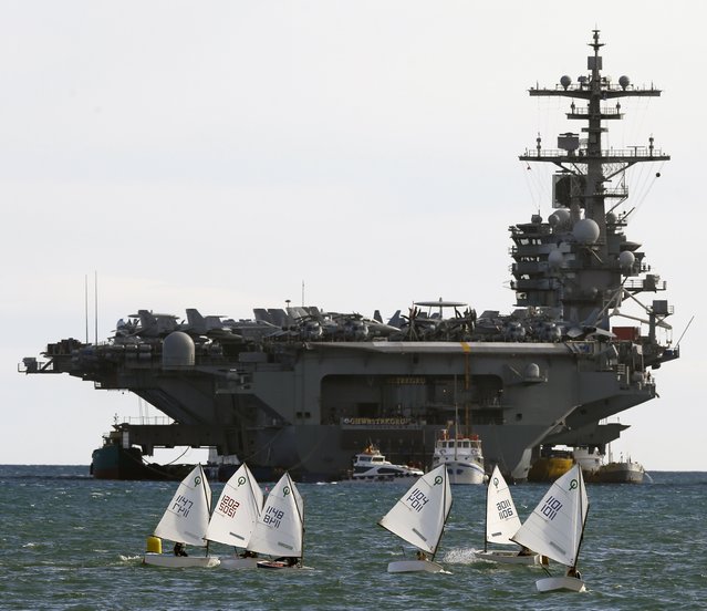People train in sail boats in the waters of the Saronic Gulf, outside Piraeus port, near Athens, Greece, back dropped by the U.S. nuclear powered aircraft carrier USS George H.W. Bush, Wednesday, March 5, 2014. The 1,092 feet (333 meters) carrier which can carry 6,000 personnel, one of the world’s largest warships, will be deployed in the U.S. 6th Fleet's area of responsibility. (Photo by Thanassis Stavrakis/AP Photo)