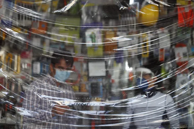 Siblings Julio Cesar, left, and Carmen Castellanos wear face masks as they wait for customers behind a protective sheet of plastic hanging over the counter in El Foquito, the hardware and electric supply store they own in the Benito Juarez district of Mexico City, Wednesday, April 15, 2020. The Castellanos family used their hardware skills and supplies to hand-make more than 100 face shields which they donated to medical staff at two hospitals in the capital. (Photo by Rebecca Blackwell/AP Photo)