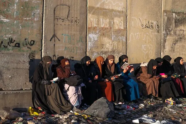 Palestinian women read the Koran as they wait in front of a concrete barrier at a checkpoint in Qalandia, in the occupied West Bank, on March 15, 2024, to cross into Jerusalem for the first Friday noon prayer of the Muslim holy fasting month of Ramadan at the Al Aqsa mosque. (Photo by Jaafar Ashtiyeh/AFP Photo)