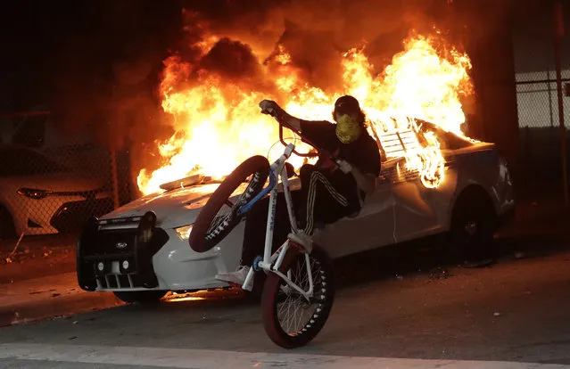 A protester on a bicycle rides past a burning police car during a demonstration next to the city of Miami Police Department, Saturday, May 30, 2020, downtown in Miami. Protests were held throughout the country over the death of George Floyd,  a black man who was killed in police custody in Minneapolis on May 25. (Photo by Wilfredo Lee/AP Photo)