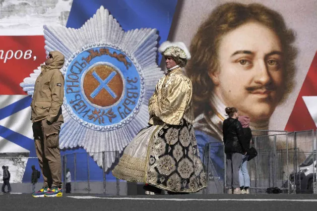 A street performer wearing a 18 century costume and earning money by posing for photos walks at the Palace Square past a portrait of Russian Tsar Peter the Great, in St. Petersburg, Russia, Friday, June 2, 2023. (Photo by Dmitri Lovetsky/AP Photo)