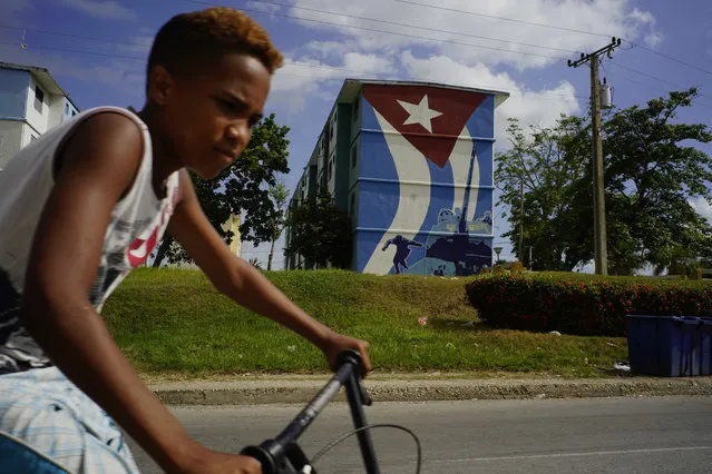In this July 25, 2018 photo, a youth rides his bike past a building covered by a mural of a Cuban flag and Fidel Castro jumping off a tank during the Bay of Pigs invasion, inspired by a photograph, in Guantanamo, Cuba, near the U.S. Guantanamo Bay naval base. Even though Fidel Castro died nearly two years ago, he lives on in the two Guantanamos. (Photo by Ramon Espinosa/AP Photo)