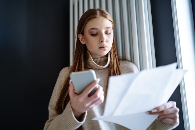 Teenage girl looking at energy bill standing by radiator heater. (Photo by Westend61/Getty Images)