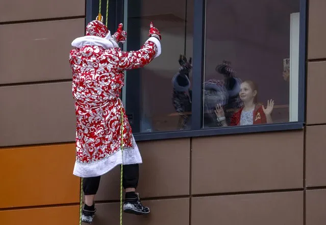 A Moscow rescuer dressed as Father Frost (or Ded Moroz, Santa Claus) greets young patients as he descends along a wall from the roof of Morozovskaya Children's hospital during a charity landing party of Fathers Frost organized for seriously-ill children ahead of the upcoming New Year and Orthodox Christmas celebrations in Moscow, Russia, 26 December 2022. Most of the children who receive medical care in the hospital are not recommended to attend crowded places and events. A team of 40 Fathers Frost accompanied by Snow Maidens visited the young patients as part of the fifth anniversary festival of good deeds “Winter in Morozovka”. (Photo by Sergei Ilnitsky/EPA/EFE)