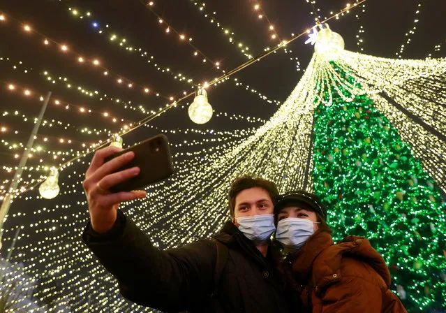 A couple wearing protective masks against COVID-19 take a selfie in a front of Christmas tree in central Kyiv, Ukraine on December 20, 2020. (Photo by Valentyn Ogirenko/Reuters)