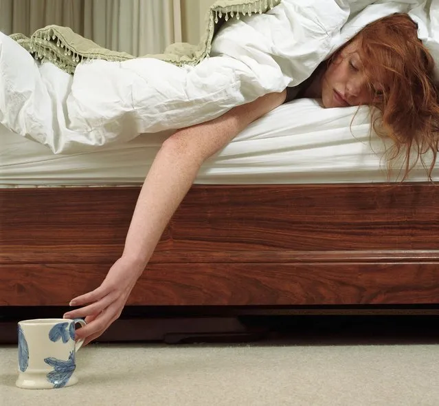 Woman lying in bed reaching for mug on carpet, eyes closed. (Photo by Alex and Laila/Getty Images)