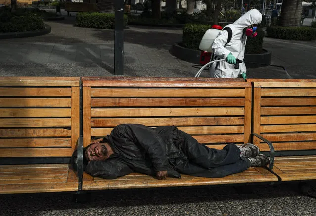A city worker sprays disinfectant while a man sleeps on a bench in the Plaza de Armas in Santiago, Chile, Wednesday, April 15, 2020.  A woman who has tested positive for the new coronavirus broke her mandatory quarantine on Wednesday and visited the Plaza de Armas to “stretch her legs” she said. The 49-year-old woman, a Peruvian national and resident in Chile, was detained by police while resting on a bench in the plaza, accompanied by her son and his girlfriend. (Photo by Esteban Felix/AP Photo)