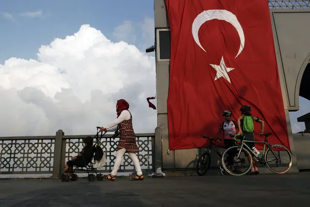 A woman passes with her child by two cyclist as they stand by a giant Turkey flag at the Galata bridge in Istanbul, on Thursday, August 4, 2016. The Turkish government characterizes the movement of Fethullah Gulen, who lives in self-imposed exile in Pennsylvania, as a terrorist organization. (Photo by Petros Karadjias/AP Photo)