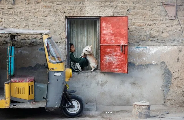 Sania, 10, plays with a dog, named Dugoo, while sitting on a window of her family home in Karachi, Pakistan on January 19, 2022. (Photo by Akhtar Soomro/Reuters)