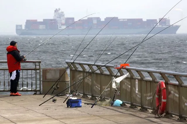 A man fishes as a container ship passes by in Kawasaki, Monday, April 11, 2022. (Photo by Koji Sasahara/AP Photo)