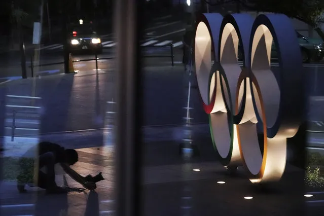 A photographer adjusting a camera to shoot the Olympic rings is reflected on a glass window near the New National Stadium Wednesday, July 22, 2020, in Tokyo. The postponed Tokyo Olympics have again reached the one-year-to-go mark. But the celebration is small this time with more questions than answers about how the Olympics can happen in the middle of a pandemic. That was before COVID-19 postponed the Olympics and pushed back the opening to July 23, 2021. (Photo by Eugene Hoshiko/AP Photo)