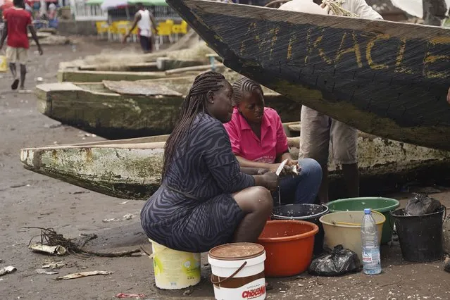 Fishmongers sit next to boats as they prepare freshly caught fish to be smoked on Limbe beach, Cameroon, on April 12, 2022. In recent years, Cameroon has emerged as one of several go-to countries for the widely criticized “flags of convenience” system, under which foreign companies can register their ships even though there is no link between the vessel and the nation whose flag it flies. But experts say weak oversight and enforcement of fishing fleets undermines global attempts to sustainably manage fisheries and threatens the livelihoods of millions of people in regions like West Africa. (Photo by Grace Ekpu/AP Photo)