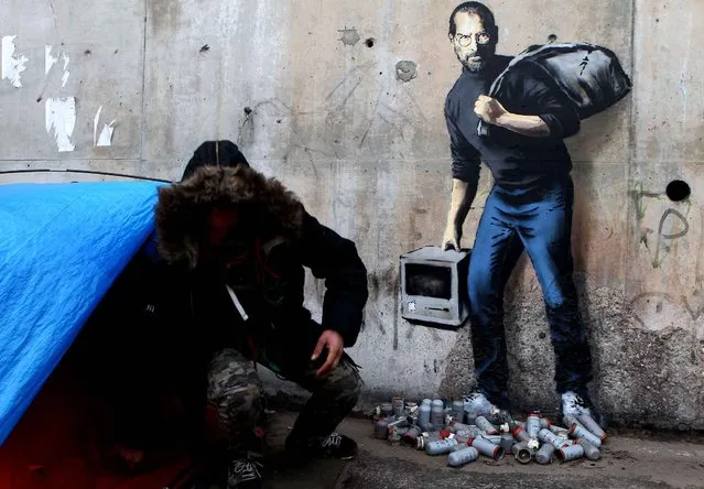 A painting by English graffiti artist Banksy is seen at the entrance of the Calais refugee camp in France, Saturday, December 12, 2015. Street artist Banksy has taken on the migrant crisis in a new mural at a migrant camp in France. The elusive graffiti artist has depicted the late Apple guru Steve Jobs – whose biological father was from Syria – carrying a black garbage bag and an early model of the Macintosh computer. (Photo by Michel Spingler/AP Photo)
