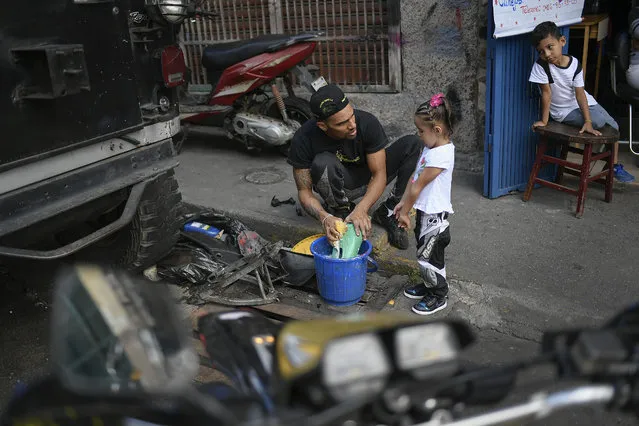 Motorcycle stuntman Pedro Aldana cleans motorcycle parts with his daughter Alanis as his young neighbor and admirer, 6-year-old Milan Sandoval Ramos, watches from a chair outside their home in the Catia neighborhood of Caracas, Venezuela, Thursday, January 21, 2021. Of Aldana's two children, 4-year-old Alanis is the only one interested in his work, who he plans to teach her how to ride a motorcycle. (Photo by Matias Delacroix/AP Photo)