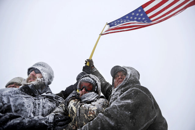 Military veterans huddle together to hold a United States flag against strong winds during a march to a closed bridge outside the Oceti Sakowin camp where people have gathered to protest the Dakota Access oil pipeline in Cannon Ball, N.D., Monday, December 5, 2016. (Photo by David Goldman/AP Photo)
