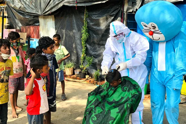 A barber wearing protective gear gives a haircut to a child next to a man dressed as comic character Doraemon during an event organised by a non-governmental organization (NGO) in a slum as the lockdown continues amid Covid-19 coronavirus pandemic in Mumbai on May 24, 2021. (Photo by Sujit Jaiswal/AFP Photo)