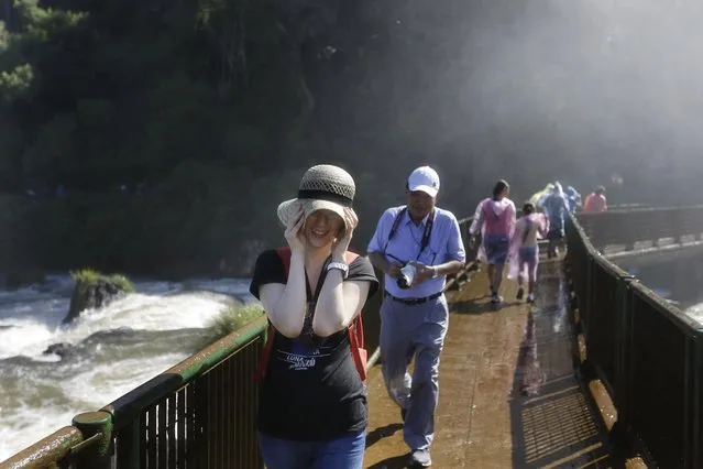 In this March 15, 2015 photo, a Japanese tourist holds on to her hat as she walks in the spray from the Iguazu Falls in Brazil. Spray from the falls douses the nearby viewing areas where some tourists don rain ponchos while others take off their shirts and dance and hug in the drenching mist. (Photo by Jorge Saenz/AP Photo)