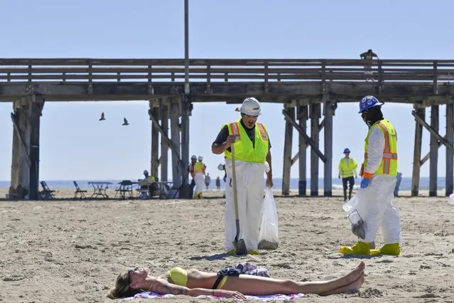 Workers clean oil from the sand, south of the pier, near a woman lying out in the sun, in Newport Beach, Calif., Tuesday, October 5, 2021. A leak in an oil pipeline caused a spill off the coast of Southern California, sending about 126,000 gallons of oil into the ocean, some ending up on beaches in Orange County. (Photo by Jeff Gritchen/The Orange County Register via AP Photo)