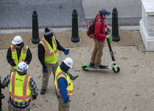 In this December 5, 2018, photo a rider maneuvers a Lime brand electric scooter on a sidewalk past workers on Capitol Hill in Washington. Electric scooters are overtaking station-based bicycles as the most popular form of shared transportation outside transit and cars. (Photo by J. Scott Applewhite/AP Photo)