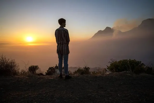 A Capetonian watches the sun rise from the top of Signal Hill as smoke engulfs the city of Cape Town, South Africa, Tuesday April 20, 2021. A massive fire spreading on the slopes of the city's famed Table Mountain, at right, is kept under control as firemen and helicopters take advantage of the low winds to contain the blaze. (Photo by Jerome Delay/AP Photo)
