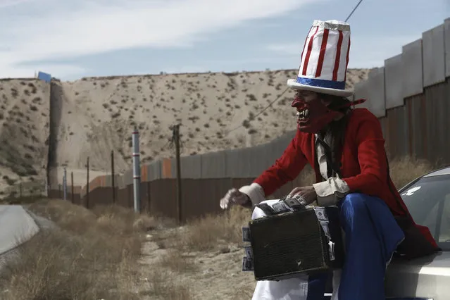 In this Sunday, February 26, 2017 photo, a protester dressed as a diabolical version of Uncle Sam holds a suitcase full of money at the U.S. border fence in Ciudad Juarez, Mexico. A group of about 30 protestors gathered to paint slogans on the border wall and stage a performance mocking the relationship between Presidents Donald Trump and Enrique Pena Nieto. (Photo by Christian Torres/AP Photo)