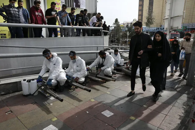 Firefighters prepare their fogging machines to disinfect a street against the new coronavirus as some people watch and pedestrians walk, in western Tehran, Iran, Friday, March 13, 2020. The new coronavirus outbreak has reached Iran's top officials, with its senior vice president, Cabinet ministers, members of parliament, Revolutionary Guard members and Health Ministry officials among those infected. The vast majority of people recover from the new coronavirus. According to the World Health Organization, most people recover in about two to six weeks, depending on the severity of the illness. (Photo by Vahid Salemi/AP Photo)