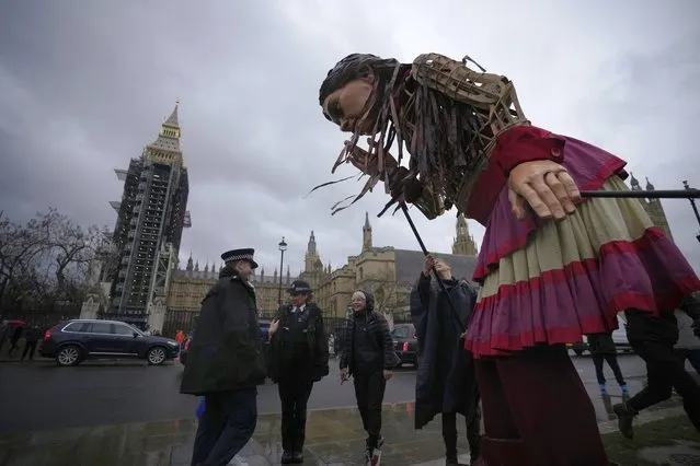 “Little Amal”, a giant puppet (3.5 meter-tall) of a young Syrian refugee girl is leaned down towards police officers on Parliament Square, backdropped by the scaffolded Elizabeth Tower, known as Big Ben, and the Houses of Parliament, in London, to coincide with the Nationality and Borders Bill being debated today in the House of Commons, Tuesday, December 7, 2021. (Photo by Matt Dunham/AP Photo)