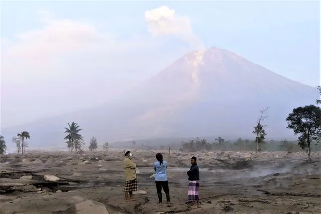 Villagers stand on an area covered in volcanic ash as Mount Semeru looms in the background in Kajar Kuning village in Lumajang, East Java, Indonesia, Monday, December 5, 2022. Indonesia's highest volcano on its most densely populated island released searing gas clouds and rivers of lava Sunday in its latest eruption. (Photo by Imanuel Yoga/AP Photo)