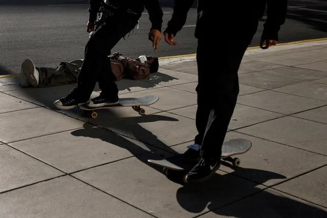 A man lays on the sidewalk wearing a protective face mask as skateboarders ride by in downtown Los Angeles, California, U.S., January 30, 2022. (Photo by Shannon Stapleton/Reuters)