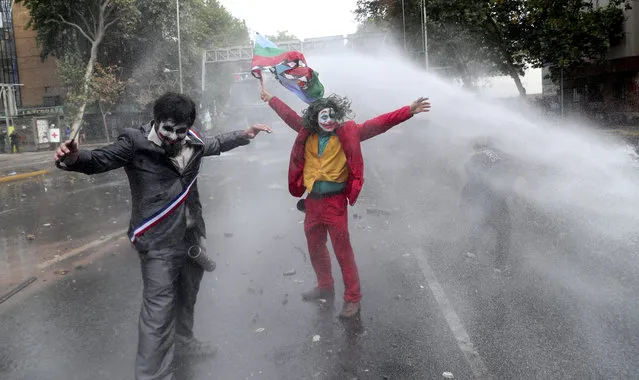 Men dressed as clowns, one dressed as the the movie character “The Joker” flying a Mapuche indigenous flag, are sprayed by a police water cannon during an anti-government protest in Santiago, Chile, Monday, November 4, 2019. Chile has been facing weeks of unrest, triggered by a relatively minor increase in subway fares. The protests have shaken a nation noted for economic stability over the past decades, which has seen steadily declining poverty despite persistent high rates of inequality. (Photo by Esteban Felix/AP Photo)