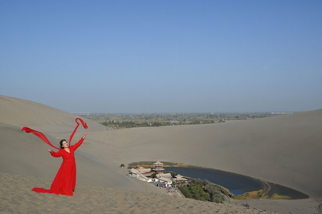 A woman has her picture taken with the Crescent Moon Spring (R) at the Mingsha mountain and Crescent Moon Spring scenic area in Dunhuang, Jiuquan, northwestern China's Gansu province on October 19, 2025. (Photo by Adek Berry/AFP Photo)