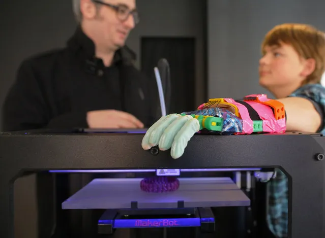 Twelve-year-old Leon McCarthy (R) rests his prosthetic hand on a MarkerBot Replicator 2 Desktop 3D Printer, while talking to the company's CEO Bre Pettis, at the new MakerBot store in Boston, Massachusetts November 21, 2013. McCarthy, whose prosthetic hand is made of parts printed from a MakerBot 3D printer, broke a piece of it last week while playing football, so he printed a new finger to repair it. (Photo by Brian Snyder/Reuters)