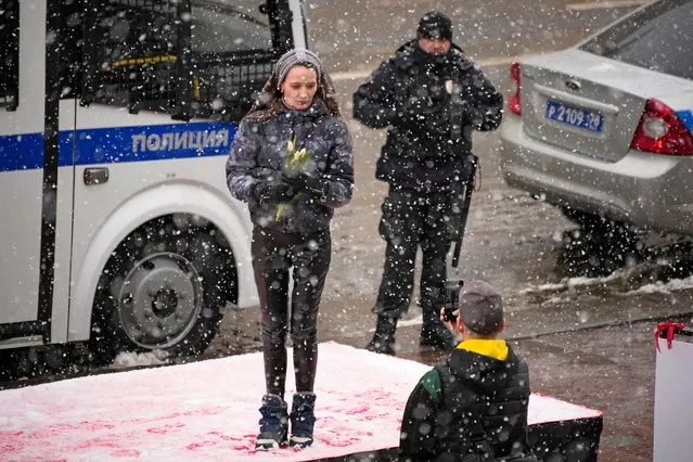 A woman poses for photos in snowfall on a podium installed on International Women's Day in central St. Petersburg, Russia, Wednesday, March 8, 2023, with a police officer in the background. International Women's Day on March 8 is an official holiday in Russia, where men give flowers and gifts to female relatives, friends and colleagues. (Photo by Dmitri Lovetsky/AP Photo)